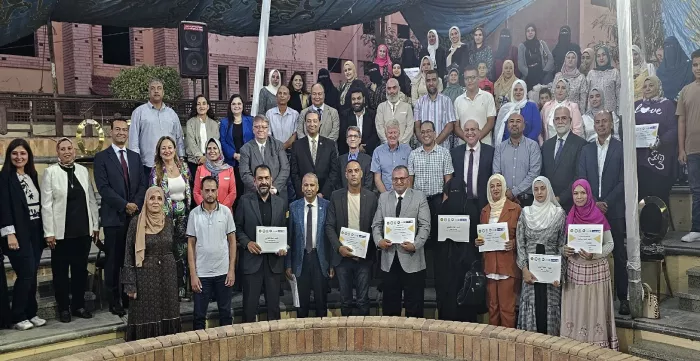 A large group of people, both men and women, pose for a photo outdoors. Some hold certificates. They stand on steps with a building and greenery in the background.