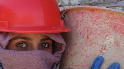 A woman with a red hard hat, holding bucket of concrete on her shoulder at a construction site